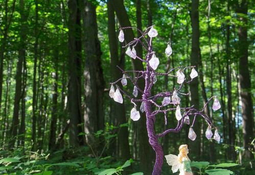 A whimsical purple tree with crystal-like leaves stands in a lush green forest, beside a small fairy figure.