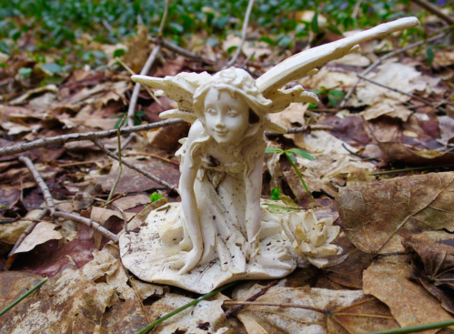 A small, white fairy figurine sits among fallen leaves and twigs, with a lotus flower at its base.