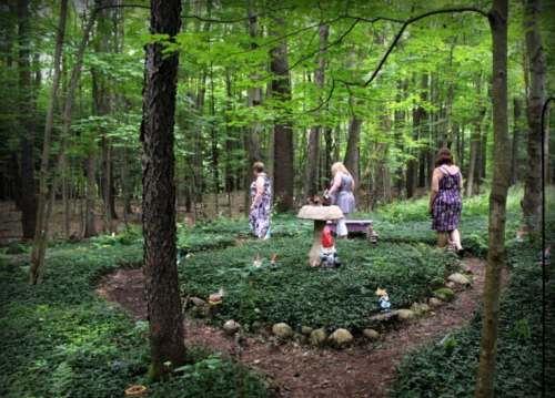 A serene forest scene with four women walking through a lush, green garden filled with small statues and a stone table.