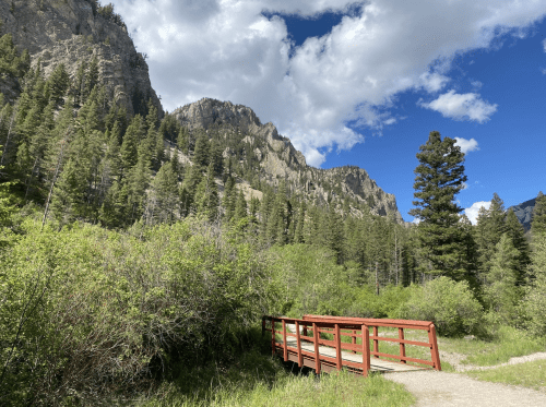 A red wooden bridge crosses a path surrounded by lush greenery and mountains under a partly cloudy sky.