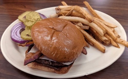 A burger with bacon, lettuce, and onion on a bun, served with pickles and a side of golden fries.
