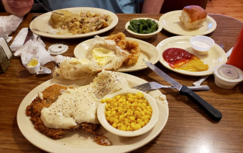 A plate of fried chicken with gravy, corn, green beans, rolls, and condiments on a wooden table.