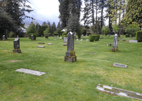 A peaceful cemetery with several gravestones set in a grassy area surrounded by trees and shrubs.