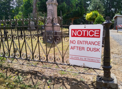 A sign on a fence reads "NOTICE: NO ENTRANCE AFTER DUSK. VIOLATORS WILL BE PROSECUTED," near a graveyard.