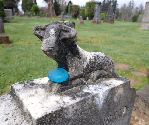 A weathered stone lamb statue rests on a grave, with a blue egg placed in front of it, set in a grassy cemetery.