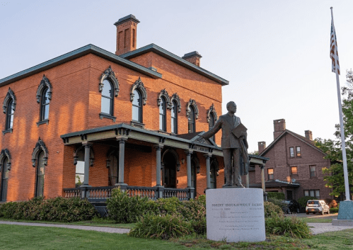 A historic brick building with a statue of Robert Brockett Johnson in front, surrounded by greenery and an American flag.