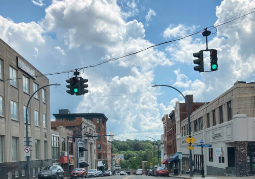 A street view with green traffic lights, buildings on either side, and a blue sky with fluffy clouds above.