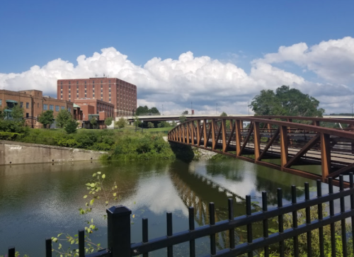 A metal bridge spans a calm river, with buildings and fluffy clouds in the background under a blue sky.