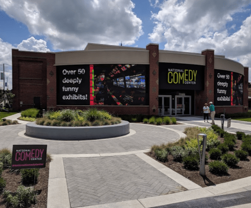 Exterior of the National Comedy Center with large signs promoting exhibits, surrounded by greenery and a blue sky.