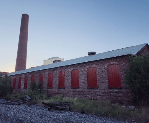 Abandoned brick building with red boarded windows and a tall chimney, set against a twilight sky.