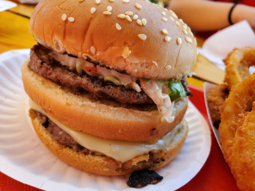 Close-up of a sesame seed bun burger with two beef patties, melted cheese, and toppings, served on a paper plate.