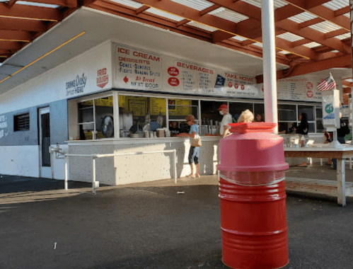 A retro ice cream stand with a menu, people ordering, and a red barrel in the foreground. Bright, sunny day.
