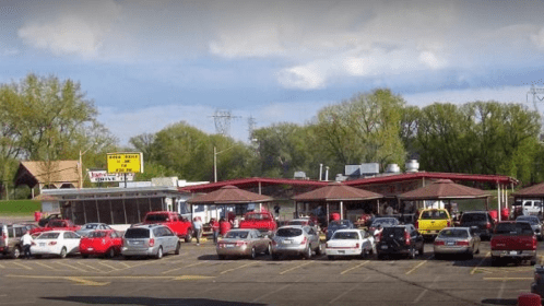 A busy outdoor food stand with a red roof, surrounded by parked cars and green trees under a cloudy sky.