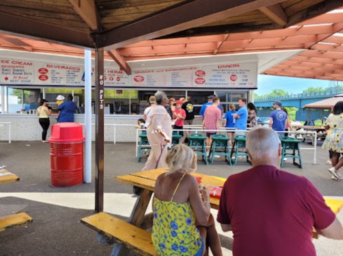 A busy outdoor food stand with people ordering and enjoying meals at picnic tables under a shaded area.