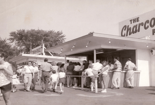 A vintage black-and-white photo of a busy food stand with people lining up and walking by in a lively outdoor setting.