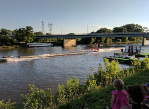 A river scene with a bridge overhead, boats on the water, and spectators on the grassy bank enjoying the view.