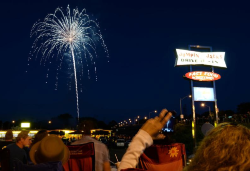 Fireworks illuminate the night sky above a drive-in restaurant, with people sitting in lawn chairs below.