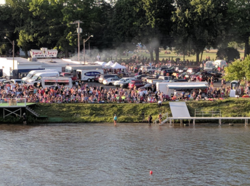 A large crowd gathers by a river, with food trucks and tents in the background, under a clear sky.