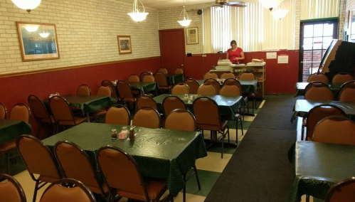 A nearly empty restaurant with green tablecloths, brown chairs, and a staff member preparing food in the background.