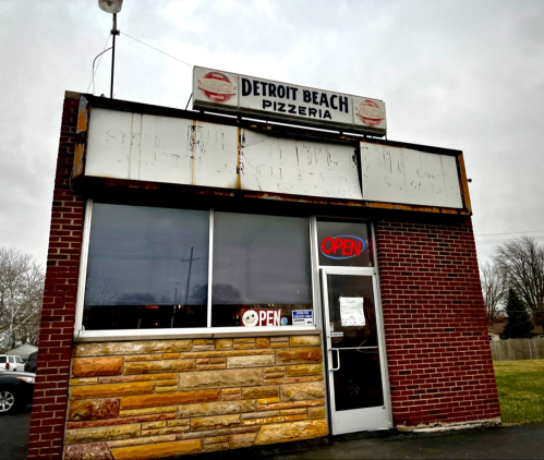 Exterior of Detroit Beach Pizzeria with a brick facade and an "OPEN" sign in the window on a cloudy day.