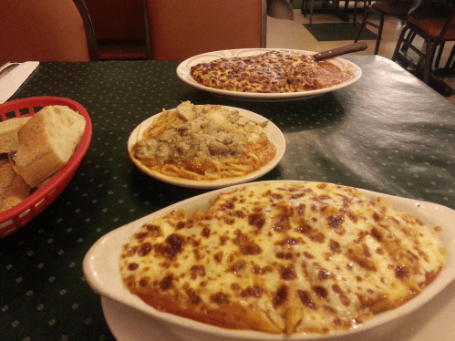 A table with three Italian dishes: baked pasta with cheese, spaghetti with meatballs, and a side of bread.