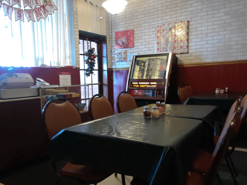 Cozy diner interior with green tables, brown chairs, and a vintage jukebox against a brick wall. Holiday decor visible.