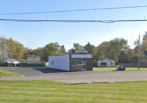 A small, vacant building on a grassy lot, surrounded by trees and utility poles under a clear blue sky.