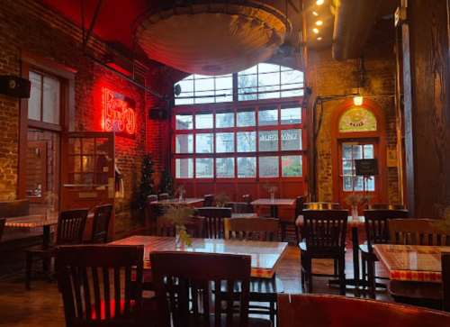 Cozy restaurant interior with wooden tables, chairs, and large windows, featuring warm lighting and exposed brick walls.
