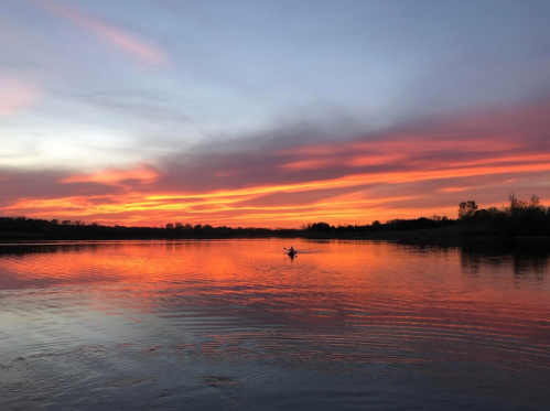 A serene sunset over a calm lake, with vibrant orange and purple hues reflecting on the water. A lone figure kayaks.