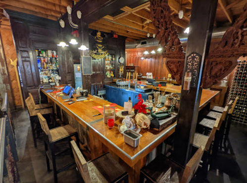 A cozy bar with wooden counters, stools, and decorative elements, featuring shelves stocked with various bottles.