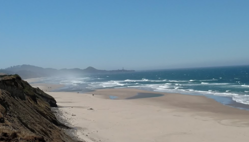 A scenic beach with gentle waves, sandy shore, and distant hills under a clear blue sky.