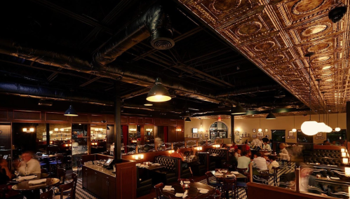A stylish restaurant interior with dark wood accents, patterned ceiling, and diners at tables. Warm lighting creates a cozy atmosphere.