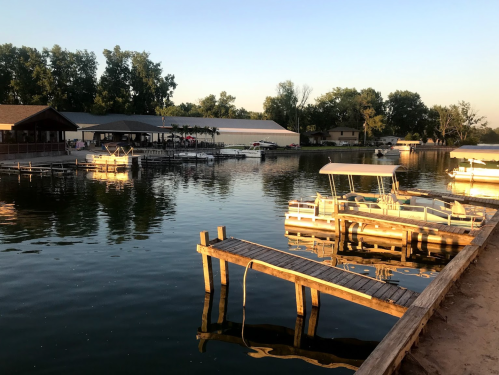 A serene lakeside scene at sunset, featuring docks and boats reflecting on calm water. Trees line the shore in the background.