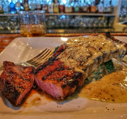 A grilled steak with a creamy sauce, served on a plate with broccoli, and a glass of whiskey in the background.