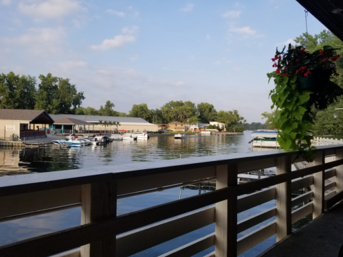 A peaceful lakeside view with boats docked, surrounded by trees and a flowered railing in the foreground.
