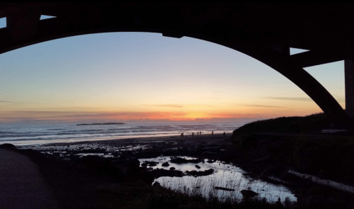 Silhouette of an arch over a beach at sunset, with people walking along the shore and reflections in the water.
