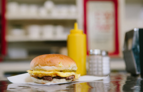 A close-up of a breakfast sandwich with egg and sausage on a paper napkin, with a bottle of mustard in the background.