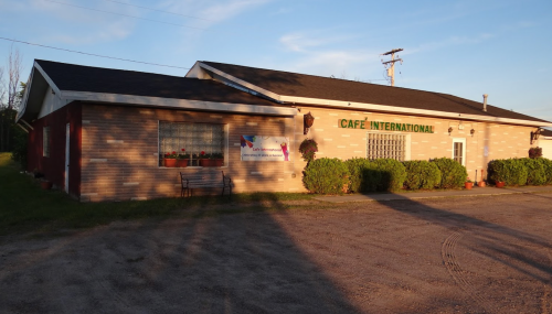 A brick building with a sign reading "Café International," surrounded by greenery and a gravel parking area.