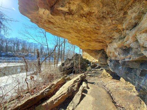 A rocky overhang beside a frozen river, surrounded by bare trees and a clear blue sky.