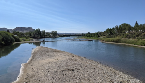 A serene river scene with sandy banks, lush greenery, and a distant bridge under a clear blue sky.