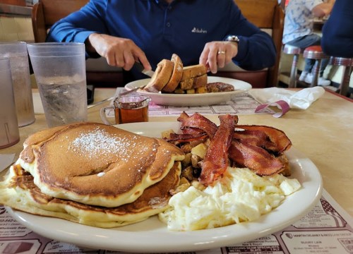 A plate of pancakes, bacon, eggs, and hash browns, with a person eating toast in the background at a diner.