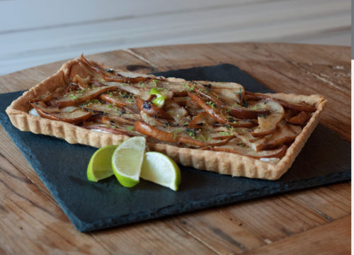 A rectangular tart topped with caramelized onions, served on a slate plate with lime wedges.