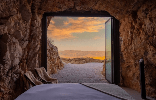 A cozy bed inside a cave, with a stunning view of a sunset through an open door.