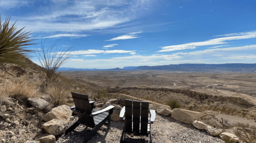 Two wooden chairs overlook a vast desert landscape under a blue sky with scattered clouds.