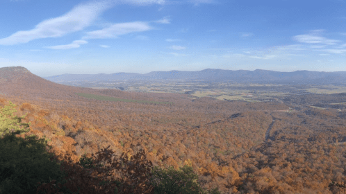 A panoramic view of a valley filled with autumn foliage, surrounded by mountains under a clear blue sky.