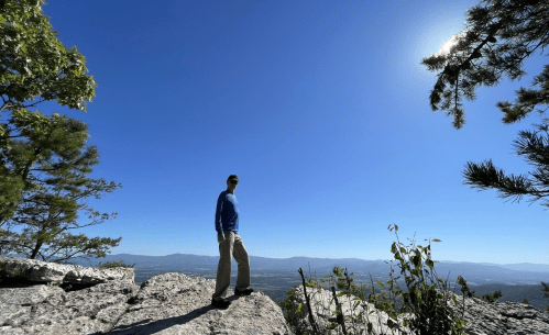 A person stands on a rocky outcrop, overlooking a vast landscape under a clear blue sky with the sun shining.
