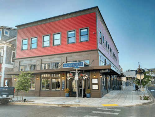 A colorful restaurant building with a red upper facade and large windows, featuring outdoor seating and decorative plants.