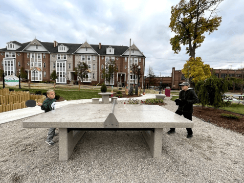 A child and an adult play table tennis at a park with modern buildings in the background.