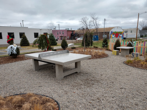 A ping pong table in a park surrounded by gravel, trees, and colorful fences, with buildings in the background.