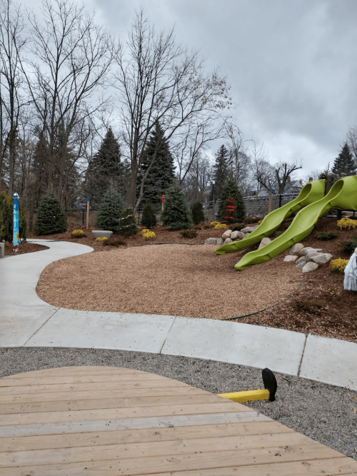 A playground area with a green slide, surrounded by trees and landscaped with mulch and rocks under a cloudy sky.